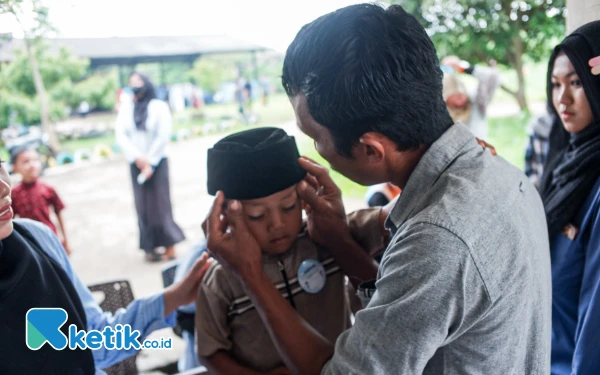 Thumbnail Seorang ayah tengah membisikkan kata-kata penenang kepada anaknya yang hendak dikhitan. Mendengar tangisan dari ruang khitan, anak-anak yang menunggu di luar menjadi merasa tegang dan ketakutan. (Foto: Wisnu Akbar Prabowo/Ketik.co.id)